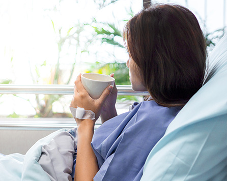 Young woman sat up in a hospital bed, looking out of the window with a cup of tea