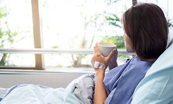 Lady in hospital bed. She is looking out the window and drinking a hot drink.